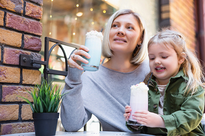 smiling girl holding glass of creamy milkshake near little daughter in street cafe