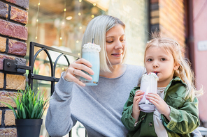 little girl drinking tasty milkshake near smiling mom in street cafe