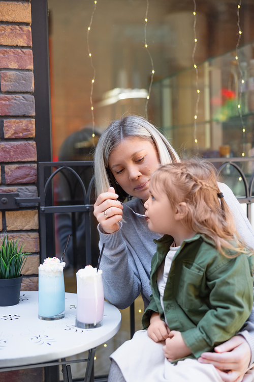 happy woman feeding daughter with milk dessert in cafe on urban street