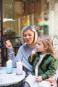 smiling woman holding spoon near daughter and tasty milkshake in street cafe