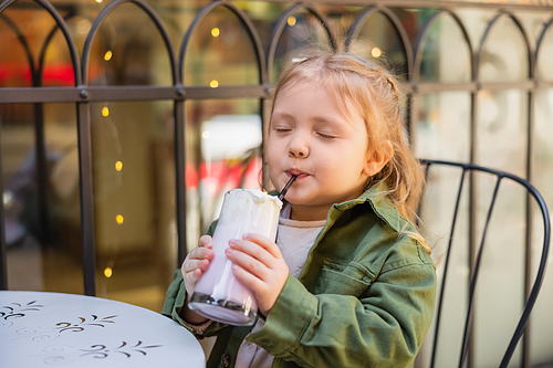 pleased girl with closed eyes drinking milkshake in street cafe