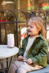smiling child looking at glass with milkshake while sitting in street cafe