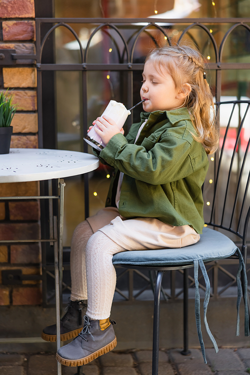 full length view of girl sitting on chair in street cafe and drinking milkshake