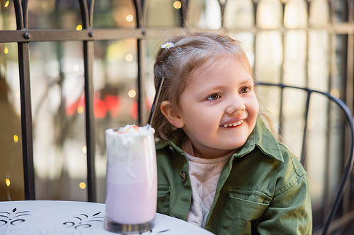 cheerful child looking away near blurred glass of milkshake outdoors