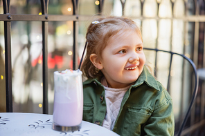 happy girl looking away near blurred glass of milkshake in street cafe