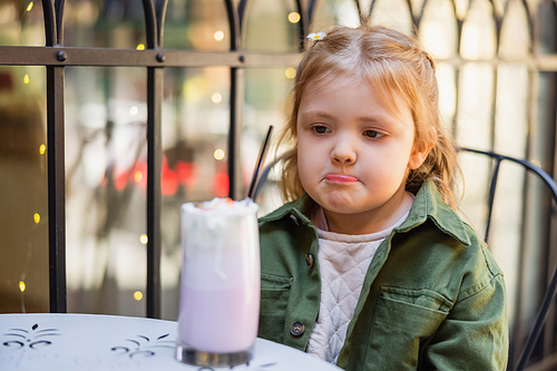 girl pouting lips while looking at blurred milkshake in street cafe
