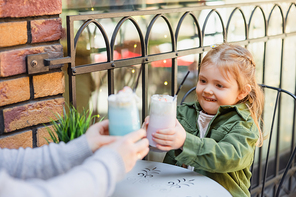 cheerful girl toasting with glasses of milkshake with blurred mom in street cafe