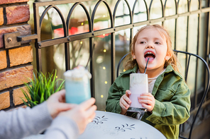 child with open mouth holding glass of milk dessert near blurred mom in street cafe