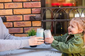 happy girl toasting with glasses of milkshake while sitting with mother in street cafe