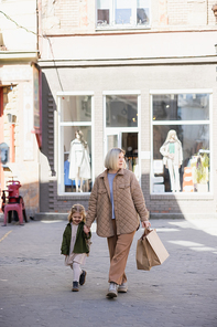 full length view of stylish woman walking with happy daughter and shopping bags outdoors
