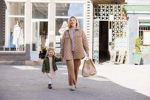smiling woman with shopping bags looking at camera while walking with child on street