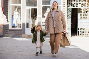 cheerful mother and daughter in trendy clothes walking with shopping bags in city