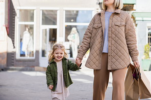 woman in spring outfit holding shopping bags while walking with daughter on urban street