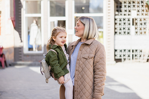 blonde woman in trendy clothes holding smiling child on city street