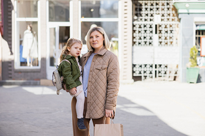 positive and trendy woman with shopping bags looking away while holding daughter outdoors