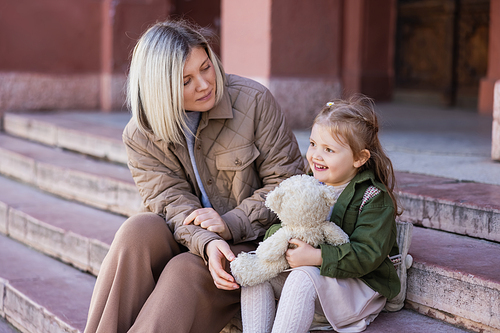 blonde woman looking at happy daughter sitting on stairs with teddy bear