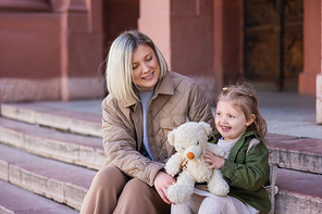 trendy and happy mother and child sitting on city street with teddy bear