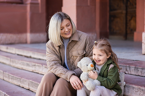 joyful child sitting on stairs with mother and teddy bear