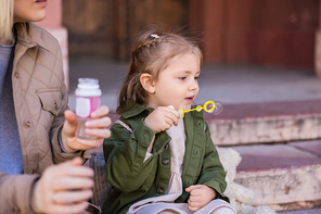 child blowing soap bubbles near blurred mother outdoors