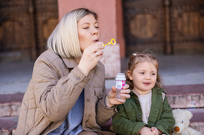 stylish woman blowing soap bubbles near smiling daughter on urban street