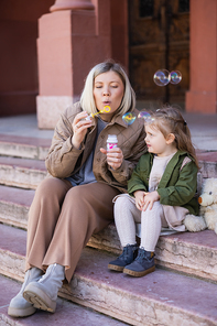 blonde woman blowing soap bubbles while sitting on stairs near daughter