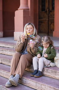 stylish woman blowing soap bubbles while sitting on street near daughter