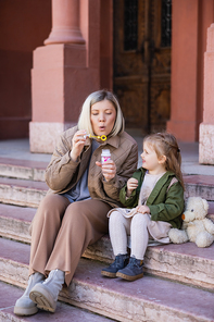 woman sitting on stairs on street and blowing soap bubbles near daughter
