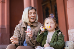 little girl blowing soap bubbles near daughter on blurred foreground