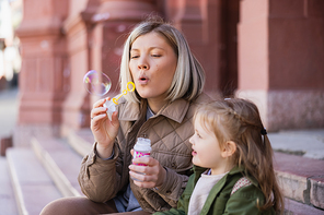 mother blowing soap bubbles near little daughter on city street