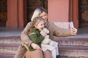 laughing girl holding teddy bear near mom taking selfie on smartphone outdoors
