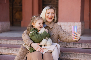 cheerful girl holding teddy bear while mom taking selfie on smartphone