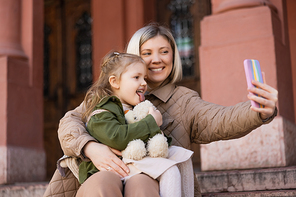 little girl with soft toy sticking out tongue near smiling mother taking selfie on mobile phone