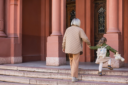 back view of woman and child with teddy bear holding hands while walking on stairs near building
