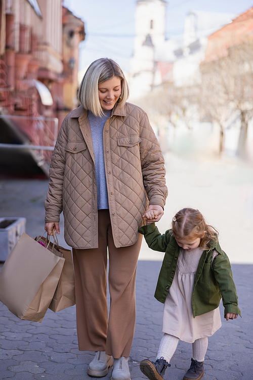 pleased woman in trendy clothes walking with shopping bags and daughter on street