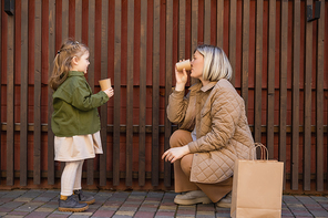 side view of mother and daughter in trendy outfit holding paper cups near wooden fence and shopping bag