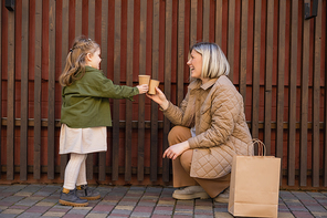 side view of happy and stylish mom and daughter toasting with paper cups near wooden fence