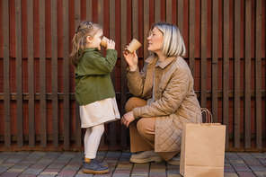 side view of mother and daughter drinking from paper cups near wooden fence and shopping bag