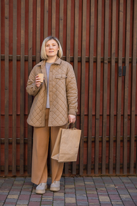 full length view of trendy woman with coffee to go and shopping bag looking at camera near wooden fence