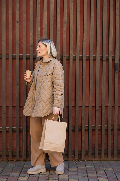 full length view of stylish woman standing with paper cup and shopping bag near wooden fence