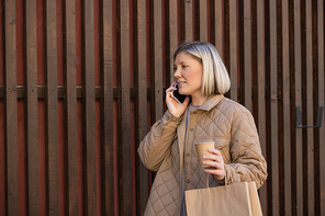 smiling blonde woman with coffee to go and shopping bag talking on smartphone near fence