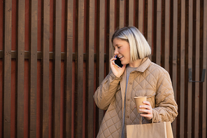 happy woman in beige jacket talking on mobile phone while holding paper cup and shopping bag