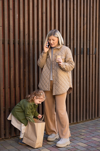 smiling woman with takeaway drink talking on cellphone near daughter looking into shopping bag