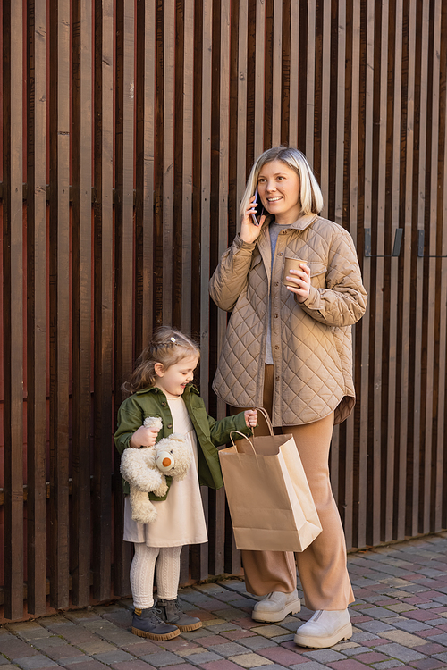 happy woman with paper cup talking on smartphone near daughter with teddy bear and shopping bag