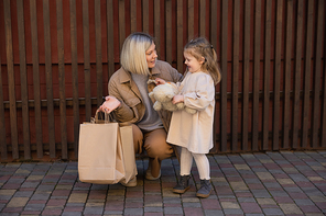 cheerful girl holding soft toy near wooden fence and mother with shopping bags