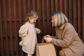 happy woman showing shopping bags to little daughter near wooden fence