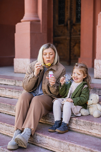 woman blowing soap bubbles near happy daughter on stairs outdoors