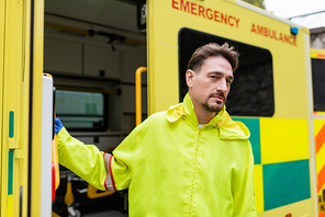 Paramedic in latex glove and jacket looking at camera near blurred ambulance vehicle