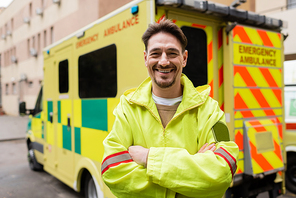 Smiling paramedic crossing arms and looking at camera near blurred ambulance vehicle outdoors