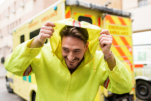 Smiling paramedic wearing hood of jacket near blurred ambulance car outdoors