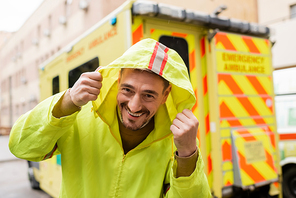 Cheerful paramedic in jacket with hood looking at camera near blurred emergency car outdoors